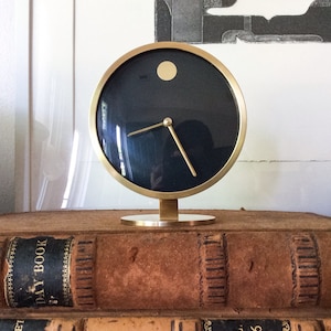 May include: A modern, round desk clock with a dark blue face and gold-colored hands and frame. The clock sits on a gold-colored base and is displayed on a stack of vintage books. The clock's face has a small gold dot at the 12 o'clock position.