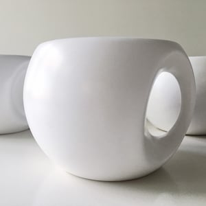 Może przedstawiać: Trzy białe, sferyczne kubki ceramiczne o unikalnym designie. Każdy kubek ma duży, okrągły uchwyt wycięty z boku. Kubki są ułożone w grupie, z jednym w ostrości, a pozostałe lekko rozmyte w tle.