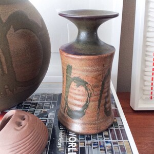 May include: A brown and green ceramic vase with a wide, flared top and a narrow base. The vase has a unique, abstract design with brown, green, and white paint.