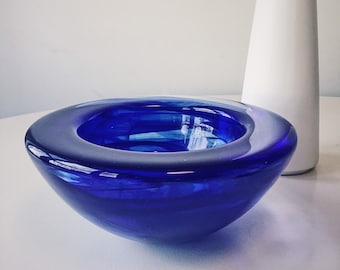 May include: A deep blue glass bowl with a wavy, organic shape. The bowl is made of thick glass and has a smooth, polished finish.