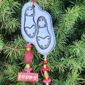 May include: A silver-colored Christmas ornament with two shoe-shaped designs. The ornament is adorned with red and yellow beads and the words "Happy Holidays" in red. It hangs from a red ribbon.