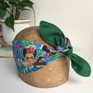 May include: A blue and green headband with a colourful print of Frida Kahlo. The headband is tied in a knot at the back.