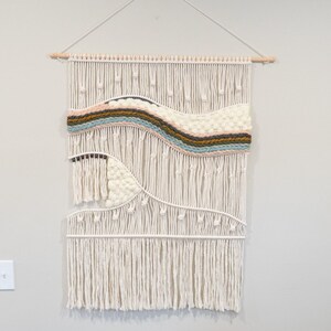 Könnte beinhalten: Ein weißer Makramee-Wandbehang mit einem gewebten Design, das ein wellenförmiges Muster in Braun-, Rosa-, Blau- und Weißtönen aufweist. Der Behang wird an einer weißen Wand über einem braunen Ledersofa mit einem weiß-grauen gemusterten Kissen gezeigt.