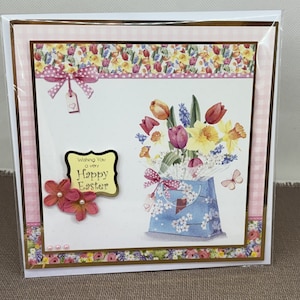 Peut inclure: Une carte de vœux faite à la main avec un fond en vichy rose et blanc. La carte présente un arrangement floral de tulipes, de jonquilles et d'autres fleurs de printemps dans un sac cadeau bleu. La carte a une bordure dorée et une étiquette jaune avec le texte "Wishing You a very Happy Easter".