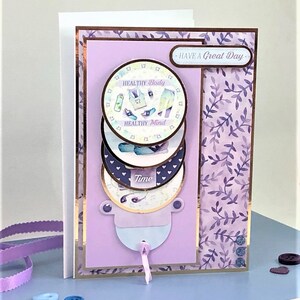 May include: A purple card with a gold border and a pull tab. The card has a layered design with three circles that say "Healthy Body", "Healthy Mind", and "Time". The card also has a purple and white floral pattern on the right side and the text "Have a Great Day" on the top right.
