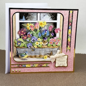 Peut inclure: Une carte rose et or avec une scène de fenêtre représentant un panier de fleurs colorées, un oiseau bleu et un papillon. La carte a un ruban doré et le texte "Wishing you a very Happy Easter".