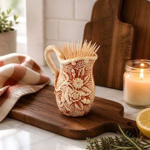 May include: A decorative toothpick holder with a handle, filled with wooden toothpicks. The ceramic holder is orange with a floral design and a cream-colored rim. It sits on a wooden cutting board, next to a lit candle and a lemon.