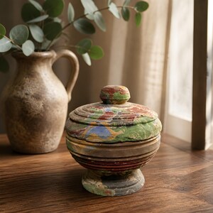 May include: A round, lidded container featuring a multi-colored marbled pattern in shades of green, red, blue, and tan. The vessel has a pedestal base, horizontal ribbed detailing along the sides, and a small, rounded knob on the center of the lid.