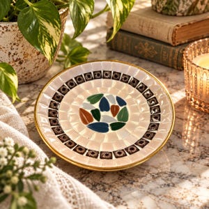 May include: A round decorative dish featuring a mosaic pattern with a gold-toned rim. The center displays leaf-shaped tiles in shades of green, blue, and brown, surrounded by concentric rings of cream-colored tiles and a band of dark patterned squares.