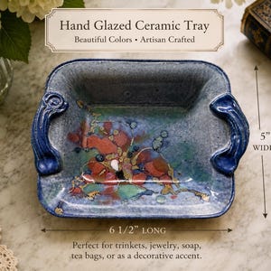 May include: A rectangular hand glazed ceramic tray with two side handles. The tray features a mottled blue and grey base with abstract red, green, and gold accents. It measures 5 inches wide and 6 1/2 inches long as indicated by the text.