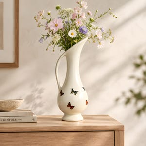 May include: A tall, cream-colored ceramic pitcher vase with a curved handle and a flared rim. The body of the vase is decorated with small, colorful butterfly illustrations in shades of orange, black, and blue, filled with a bouquet of wildflowers.