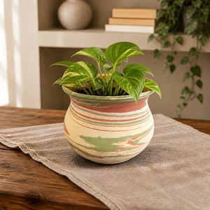 May include: A potted plant with vibrant green and yellow leaves sits in a decorative ceramic pot. The pot features a cream base with horizontal stripes of red, green, and pink. The pot is on a brown wooden table with a tan runner.