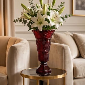 May include: A tall, ruby red glass vase filled with white lilies and green eucalyptus leaves. The vase has a detailed, textured pattern and sits on a small, round table with a gold frame. The arrangement is in a living room setting.