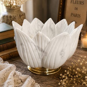 May include: A white marble bowl shaped like a blooming lotus flower with pointed petals and a smooth, veined texture. The bowl rests on a circular, polished gold-toned metal base, providing a contrast to the white stone material of the vessel.