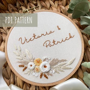 Op de afbeelding: Een geborduurde ring met de namen "Victoria & Patrick" in bruin cursief schrift. De ring is versierd met een bloemmotief met witte bloemen, gele centra en bruine stelen.
