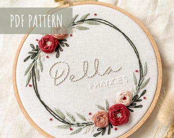 May include: Embroidered hoop art with a floral wreath design. The wreath is made of green leaves and pink and red flowers. The name "Bella Frances" is embroidered in the center of the hoop.