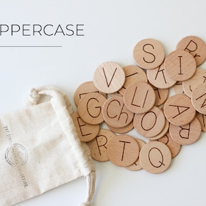 May include: A set of wooden alphabet letter tiles in a small white drawstring bag. The tiles are round and have the letters of the alphabet engraved on them. The letters are in uppercase. The bag has the text "The Simple  &  Natural Collection" printed on it.