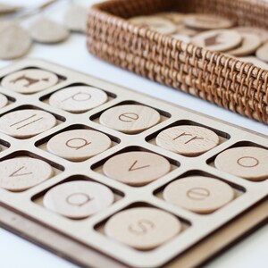 May include: A wooden tray with circular cutouts for letter tiles. The tray is filled with tiles that have letters printed on them, including "u", "v", "a", "e", "p", "r", "o", and "s".