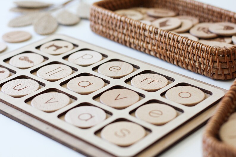 May include: A wooden tray with circular cutouts for letter tiles. The tray is filled with tiles that have letters printed on them, including "u", "v", "a", "e", "p", "r", "o", and "s".