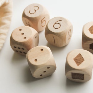 May include: Six wooden dice with engraved numbers and symbols, alongside a beige drawstring bag. The dice are light brown and have a natural wood grain. The symbols include a hexagon, a diamond, and a square.