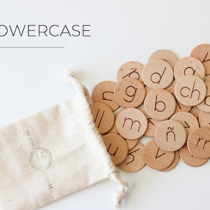 May include: A white drawstring bag with the text "The Simple Alphabet Collection" printed on it, spilling out wooden lowercase letter tiles onto a white surface. The tiles feature various letters of the alphabet, including "a", "b", "c", "d", "g", "m", "n", "r", "u", "v", "y", and "ch".