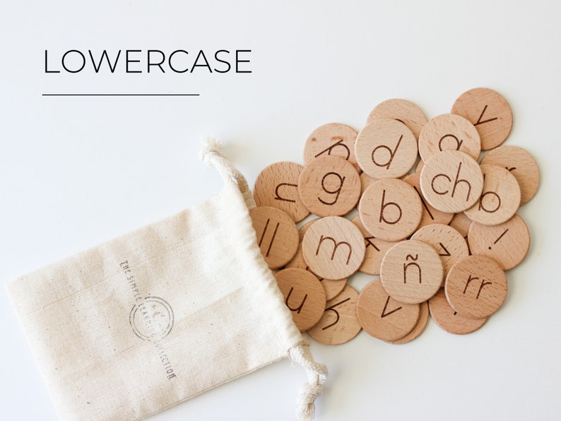 May include: A white drawstring bag with the text "The Simple Alphabet Collection" printed on it, spilling out wooden lowercase letter tiles onto a white surface. The tiles feature various letters of the alphabet, including "a", "b", "c", "d", "g", "m", "n", "r", "u", "v", "y", and "ch".