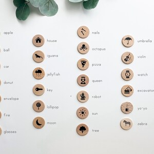 May include: A set of 24 wooden circles with black illustrations and text labels. The circles feature various objects, including an apple, house, ball, iguana, nails, umbrella, octopus, violin, pizza, car, jellyfish, watch, donut, queen, key, excavator, robot, envelope, lollipop, yo-yo, fire, sun, moon, zebra, and glasses.