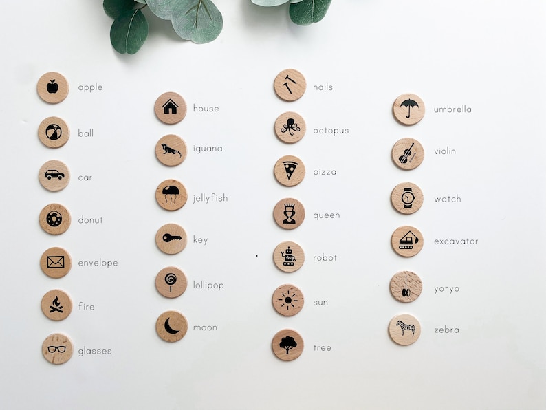 May include: A set of 24 wooden circles with black illustrations and text labels. The circles feature various objects, including an apple, house, ball, iguana, nails, umbrella, octopus, violin, pizza, car, jellyfish, watch, donut, queen, key, excavator, robot, envelope, lollipop, yo-yo, fire, sun, moon, zebra, and glasses.
