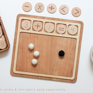 Puede incluir: Manipulador matemático de madera con un marco marrón claro y una superficie de madera clara. El marco tiene ranuras para fichas de números y símbolos. La superficie tiene tres bolas de fieltro blancas y una bola de fieltro negra. El texto "3 + / = 4" es visible en el marco.