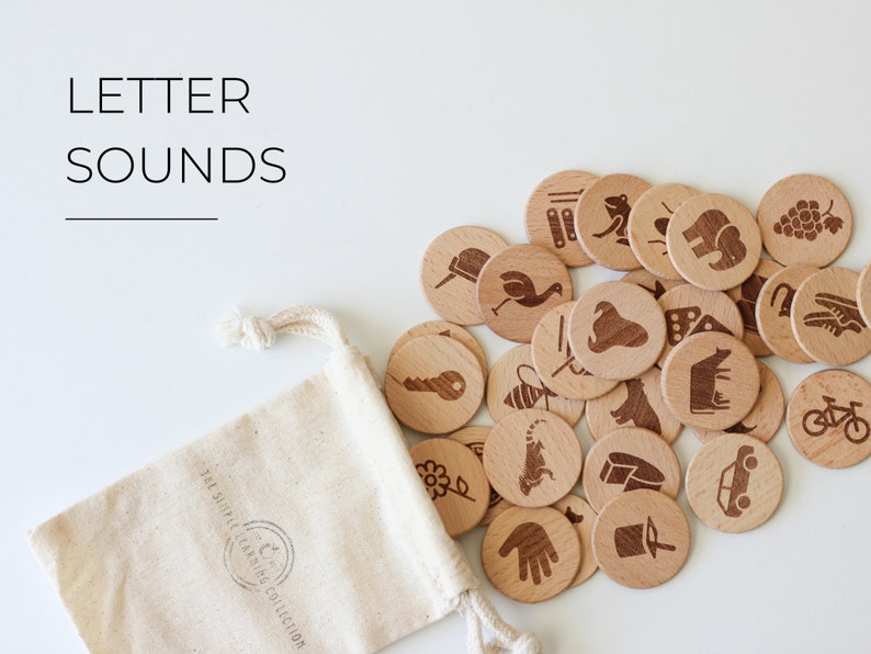 May include: A set of wooden letter sound learning tokens in a white drawstring bag. The tokens feature various images, such as a bee, a car, a cow, a frog, a hand, a key, a tiger, and a bicycle. The bag has the text "The Simple Learning Collection" printed on it.