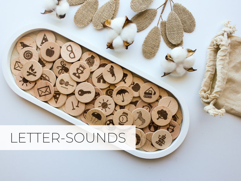 May include: Wooden tray filled with letter sound learning tokens. Each token features a different image, such as an apple, a key, and a sun. The tray is white and oval shaped. The text "LETTER-SOUNDS" is printed below the tray.