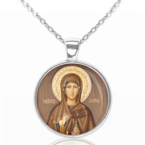 Può includere: Una collana con catena d'argento con un ciondolo rotondo che presenta un'icona religiosa di una donna con una tunica marrone e un'aureola dorata. L'icona è posizionata su uno sfondo di legno marrone con le parole "saanna" e "joanna" scritte in oro.