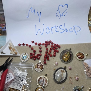 May include: A collection of jewellery-making supplies including red beads, various metal pendants with religious and portrait imagery, small loose beads, and two pairs of pliers. A white paper sign with blue handwritten text reads My Workshop.
