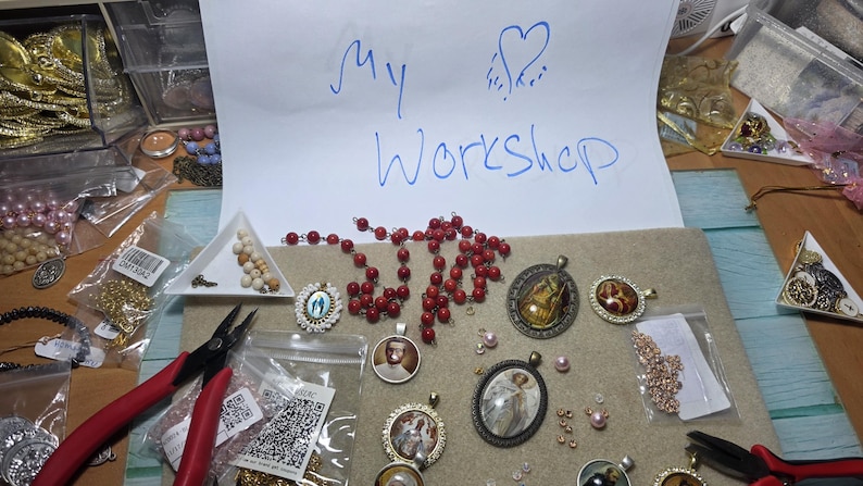 May include: A collection of jewellery-making supplies including red beads, various metal pendants with religious and portrait imagery, small loose beads, and two pairs of pliers. A white paper sign with blue handwritten text reads My Workshop.