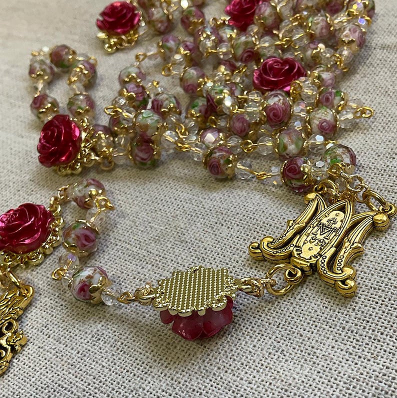 Handmade Marian Catholic rosary with pink floral glass beads, red roses, gold-tone chain and an Immaculate Mary centerpiece displayed on neutral linen.