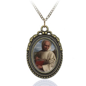May include: A gold-toned pendant necklace with a round oval-shaped frame featuring a portrait of a man in a white robe holding a red book. The frame has a decorative border with gold accents.