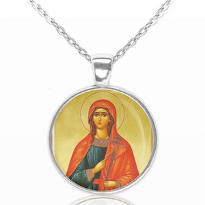 Op de afbeelding: Een zilveren ketting met een ronde hanger met een religieus icoon. Het icoon toont een vrouw in een rode mantel die een kruis vasthoudt. De achtergrond is geel en goud van kleur.