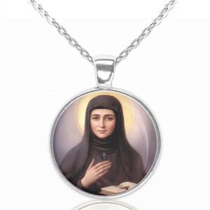 Op de afbeelding: Zilveren ketting met een ronde glazen hanger met een portret van een vrouw in een zwarte habit en een halo. De vrouw houdt een boek en een kruis vast.