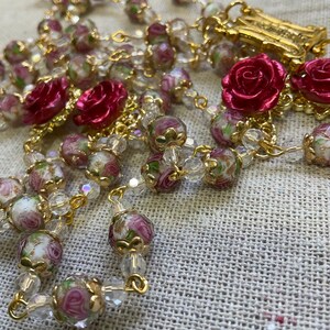 Red rose and floral glass bead Catholic rosary with gold-tone devotional components displayed on textured linen, highlighting the handmade beadwork and red, pink and gold colors.