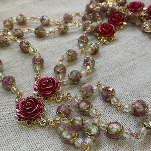 Close view of a red rose Catholic rosary with clear pink floral lampwork beads and gold-tone links arranged on light linen, showing the handmade glass bead details.