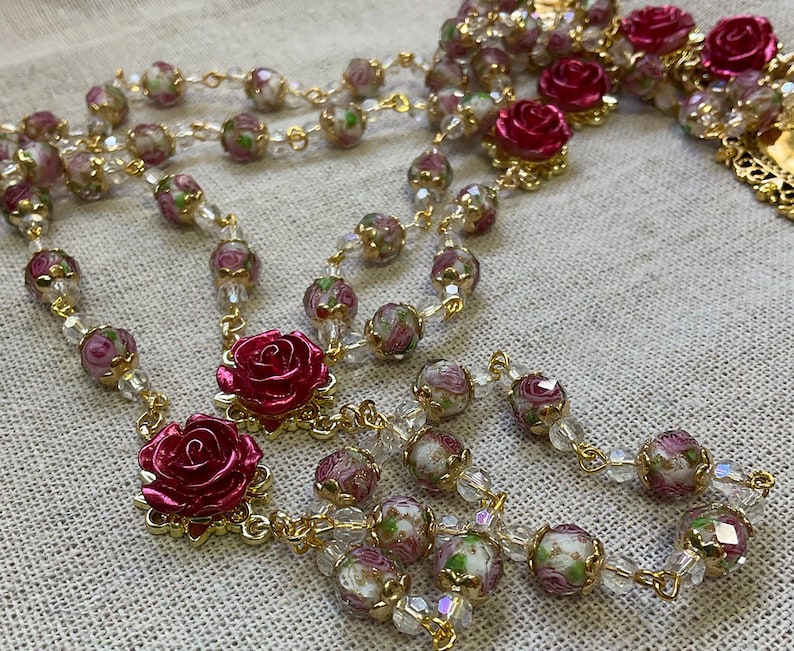 Close view of a red rose Catholic rosary with clear pink floral lampwork beads and gold-tone links arranged on light linen, showing the handmade glass bead details.