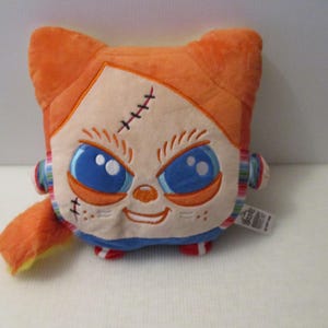 May include: A plush toy featuring a square-shaped head with bright orange hair, large blue eyes, and stitched facial details. The character has a pale face, a small orange nose, and a smiling mouth. It wears a blue shirt with multi-colored stripes.