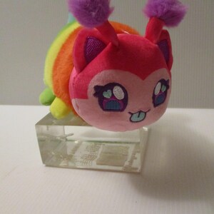 Pu&ograve; includere: Un peluche con un viso rosa, grandi occhi ricamati, una piccola lingua che sporge e due antenne viola soffici. Il corpo &egrave; a righe con i colori dell'arcobaleno, inclusi rosso, arancione, giallo e verde, appoggiato su un blocco rettangolare.