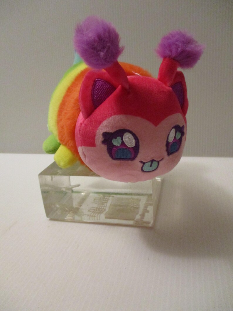 Pu&ograve; includere: Un peluche con un viso rosa, grandi occhi ricamati, una piccola lingua che sporge e due antenne viola soffici. Il corpo &egrave; a righe con i colori dell'arcobaleno, inclusi rosso, arancione, giallo e verde, appoggiato su un blocco rettangolare.
