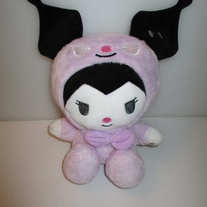 May include: A plush toy featuring a character with a white face, black hair, and a pink hood with large black ears. The character has embroidered eyes, a pink nose, and a small smile. A light pink bow is attached to the neck of the plush figure.