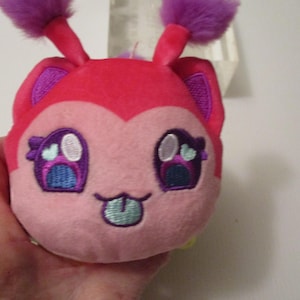 Pu&ograve; includere: Un piccolo peluche rotondo con un viso rosa acceso e rosa chiaro, dotato di due antenne viola soffici sulla parte superiore. Il viso ricamato presenta grandi occhi viola con accenti bianco e azzurro, e una piccola bocca con una lingua azzurra che sporge.