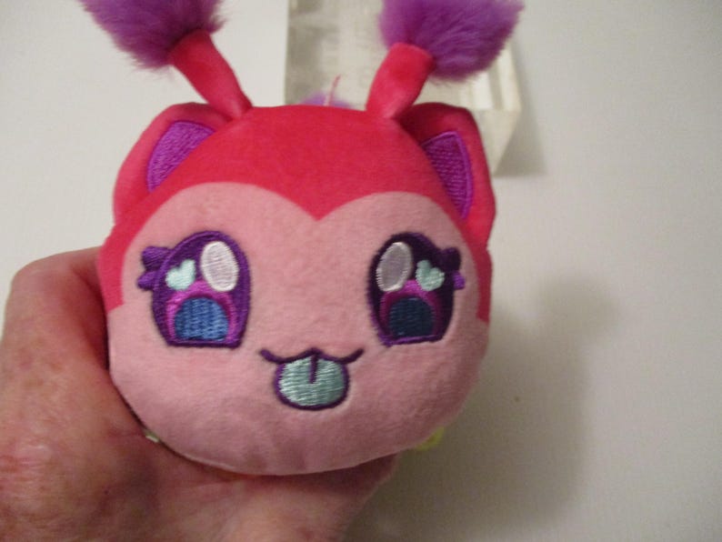 Pu&ograve; includere: Un piccolo peluche rotondo con un viso rosa acceso e rosa chiaro, dotato di due antenne viola soffici sulla parte superiore. Il viso ricamato presenta grandi occhi viola con accenti bianco e azzurro, e una piccola bocca con una lingua azzurra che sporge.