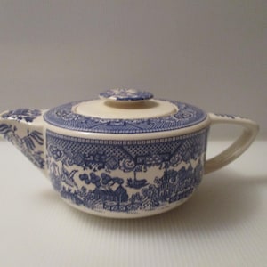 May include: A cream-colored ceramic teapot featuring a blue willow pattern. The teapot has a matching lid with a small knob, a curved handle, and a short spout. The intricate blue design depicts traditional landscape scenes with bridges and birds.