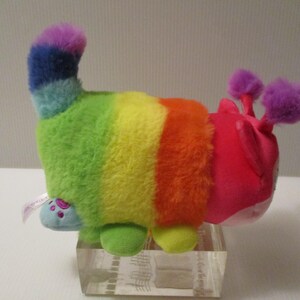 Pu&ograve; includere: Un peluche a forma di bruco con un corpo a strisce arcobaleno caratterizzato da segmenti verdi, gialli e arancioni. La testa &egrave; rosa brillante con due antenne viola soffici. Presenta una coda blu e piccoli piedi azzurri con impronte di zampe.