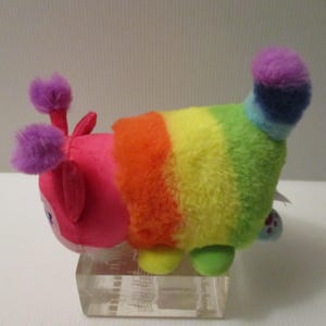 Pu&ograve; includere: Un peluche a forma di bruco con una testa rosa brillante, due antenne a pon-pon viola e un corpo caratterizzato da strisce orizzontali arancioni, gialle, verdi e blu. La coda &egrave; un ciuffo soffice viola e blu, ed &egrave; dotato di piccoli piedini.